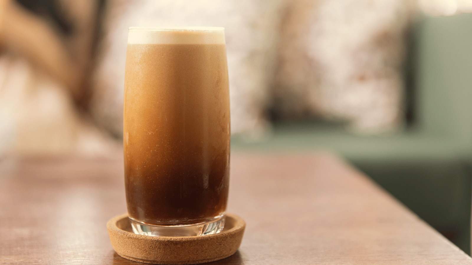 Drinking glass sitting inside a wooden coaster on a wood table. Inside the glass is nitro coffee.